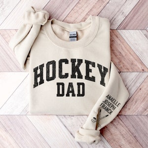 May include: A beige sweatshirt with the text "HOCKEY DAD" in black. The sleeve has the text "ANAELLE JOSEPH FRANCK" and a graphic of two hockey sticks and a puck.