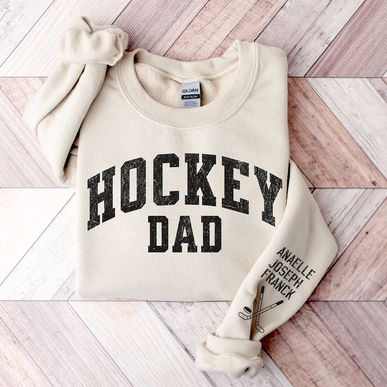May include: A beige sweatshirt with the text "HOCKEY DAD" in black. The sleeve has the text "ANAELLE JOSEPH FRANCK" and a graphic of two hockey sticks and a puck.
