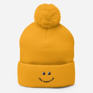 Puede incluir: Un gorro de punto amarillo con un pompón esponjoso en la parte superior. El gorro tiene una cara sonriente bordada en negro con el texto "Cool Coders Club" debajo.