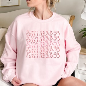 May include: Pink sweatshirt with the text "BMT NURSE" repeated in a retro, wavy font.