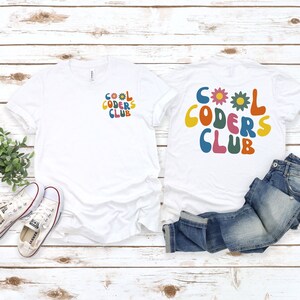 May include: Two white t-shirts with the text "Cool Coders Club" in colourful, retro-style letters. One shirt has the text on the front and the other has the text on the back. The shirts are laid out on a white wooden surface with a pair of white trainers and blue jeans.