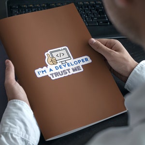 Pode incluir: Uma pasta marrom com um adesivo que diz "Sou desenvolvedor, confie em mim". O adesivo apresenta um computador de desenho animado com c&oacute;digo na tela e um polegar para cima.