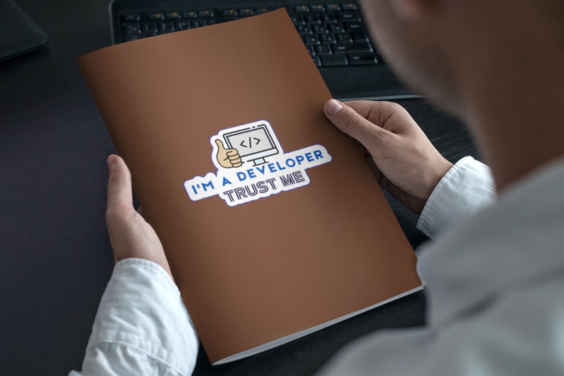 Pode incluir: Uma pasta marrom com um adesivo que diz "Sou desenvolvedor, confie em mim". O adesivo apresenta um computador de desenho animado com c&oacute;digo na tela e um polegar para cima.