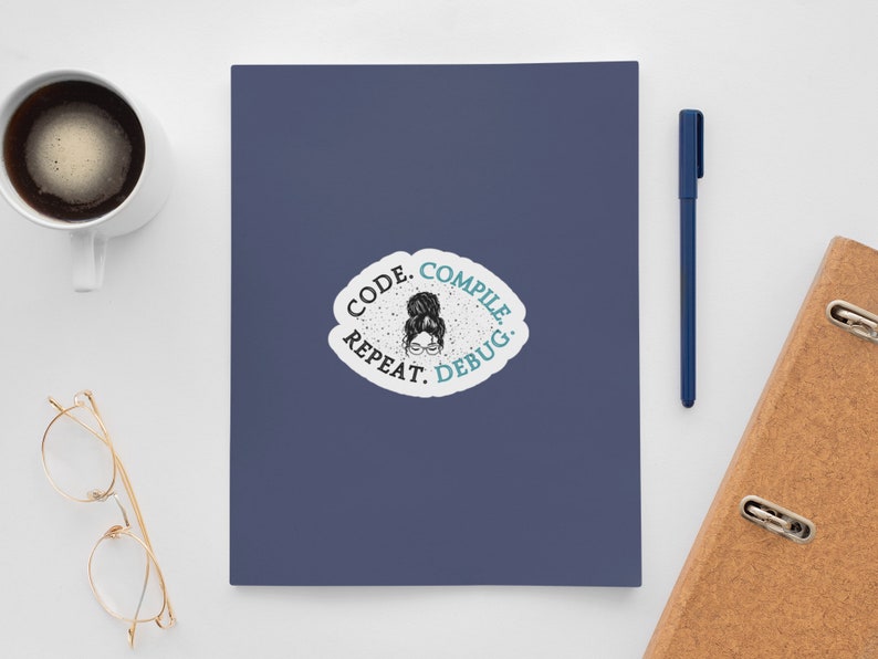 May include: A navy blue notebook with a sticker that reads "CODE. COMPILE. REPEAT. DEBUG." A cup of coffee, a pen, and a pair of glasses are also on the white surface. A brown notebook is on the right side.