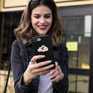 May include: A woman is holding a black smartphone with a sticker on the back. The sticker features a cartoon character with the text "Code Ninja".