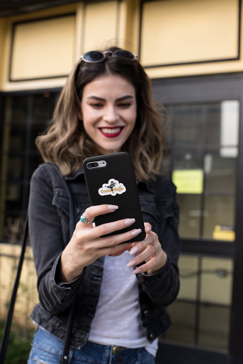 May include: A woman is holding a black smartphone with a sticker on the back. The sticker features a cartoon character with the text "Code Ninja".