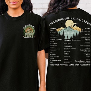 May include: Black t-shirt with a graphic of a forest scene and the text "Nature is my happy place". The back of the shirt has a list of Canadian national parks and the text "Preserving our national parks. Take only pictures, leave only footprints."