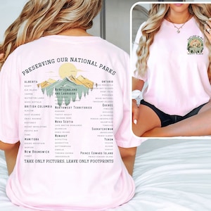 May include: Pink t-shirt with a graphic of mountains and trees, and the text "Preserving Our National Parks" and a list of national parks in Canada. The text "Take Only Pictures, Leave Only Footprints" is at the bottom of the graphic. The back of the shirt has a graphic of a mountain with a tree and the text "Nature is my happy place".
