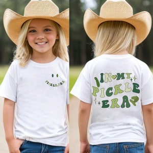 Peut inclure: Un t-shirt blanc avec un motif vert et blanc qui dit "In My Pickle Era" au dos et un smiley vert et blanc sur le devant. Le t-shirt est porté par un enfant portant un chapeau de cowboy en paille.