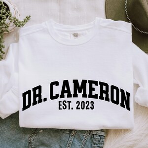 May include: White sweatshirt with black text that reads "Dr. Cameron Est. 2023".