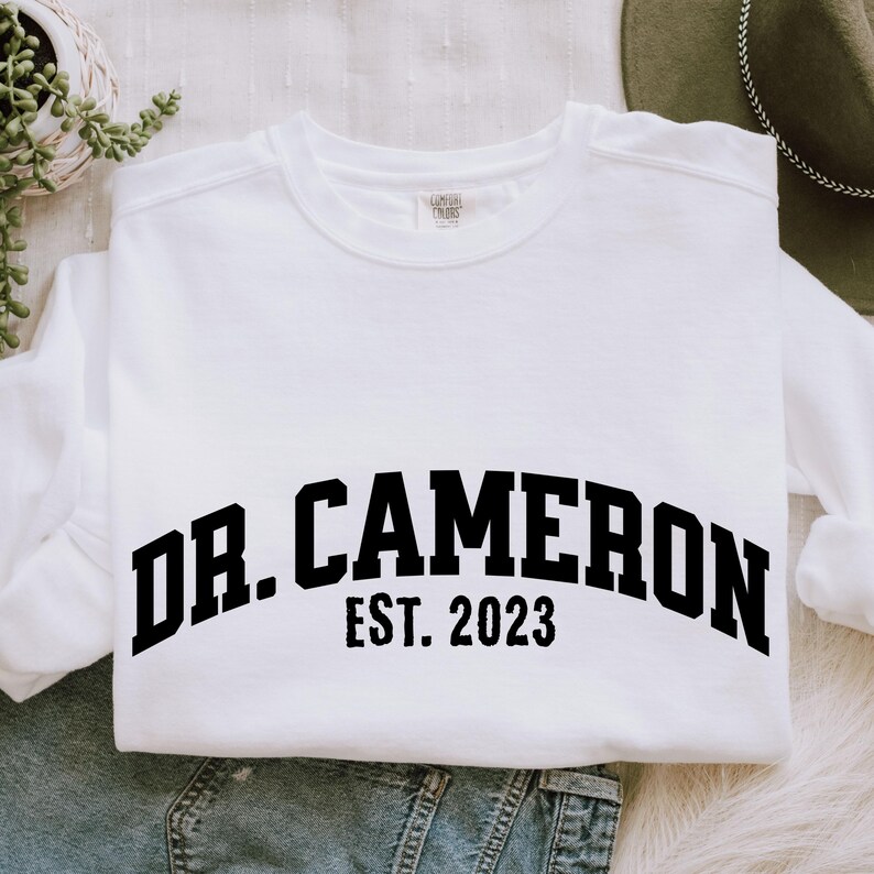 May include: White sweatshirt with black text that reads "Dr. Cameron Est. 2023".
