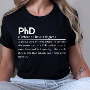 May include: Black t-shirt with white text that reads "PhD (Pretend to have a degree)" and a definition of the term.