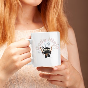 Könnte beinhalten: Eine weiße Keramiktasse mit einem Cartoon-Ninja, der Schwerter hält, und dem Text "Code Ninja" in rosa und blauen Buchstaben.