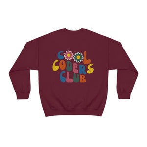 Peut inclure: Sweat-shirt bordeaux avec un graphisme r&eacute;tro qui dit "Cool Coders Club" en lettres color&eacute;es avec des fleurs.