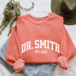 May include: A coral colored sweatshirt with white text that reads "DR. SMITH EST. 2023".