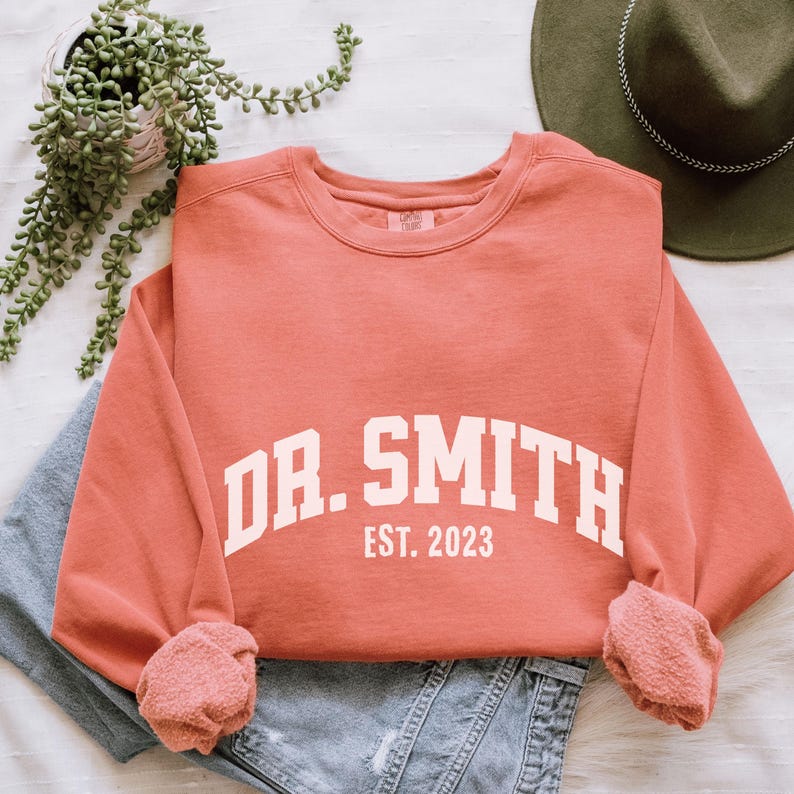 May include: A coral colored sweatshirt with white text that reads "DR. SMITH EST. 2023".