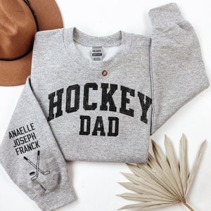 May include: Grey sweatshirt with the text "HOCKEY DAD" in black on the front. The left sleeve has the text "ANAELLE JOSEPH FRANCK" and a hockey stick and puck graphic.