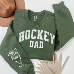 May include: Green sweatshirt with the text "HOCKEY DAD" in white on the front. The left sleeve has the text "ANAELLE JOSEPH FRANCK" and a graphic of two hockey sticks and a puck.