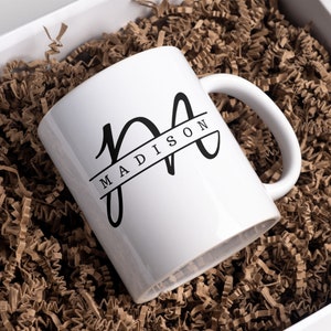 May include: White ceramic mug with a black monogram design. The monogram is a stylized "M" with the name "MADISON" written in a banner across the top of the letter.