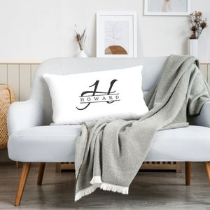 Op de afbeelding: Een wit kussen met een zwart monogram ontwerp. Het monogram is een gestileerde "H" met de naam "HOWARD" eronder.