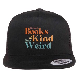 May include: Black trucker hat with the text "Read Books Be Kind Stay Weird" in orange and teal lettering. The hat has a mesh back and a curved brim. A small white tag is on the brim.