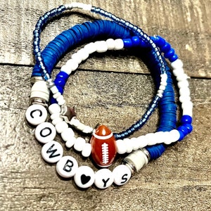 May include: A set of three bracelets with blue and white beads. The bracelets spell out "COWBOYS" with a football charm and a heart charm.