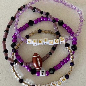 May include: A set of four beaded bracelets in shades of purple, black, and white. The bracelets feature the words "VIKINGS" and "CARSON" with a football charm and the number "7".