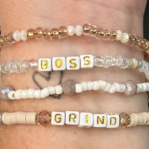 Op de afbeelding: Vier kralenarmbanden met de woorden "BOSS" en "GRIND" gespeld met witte letterkralen. De armbanden zijn op elkaar gelegd op een pols en hebben witte, beige en heldere kralen.