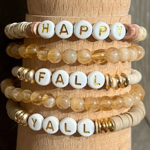 May include: A set of four bracelets with white beads and gold accents. The bracelets spell out "Happy Fall Y'all" in white letters.