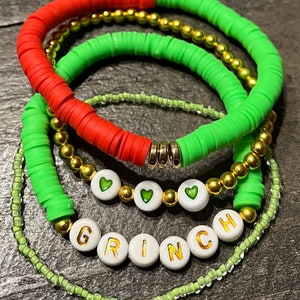 Puede incluir: Un conjunto de cinco pulseras con cuentas verdes y rojas, cuentas doradas y cuentas blancas con letras doradas que deletrean "GRINCH" y corazones verdes.