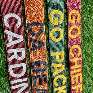 May include: Close-up of four beaded belts with team names. The belts are red, orange, green, and maroon, with white, navy, and yellow beaded lettering spelling "CARDINA", "BEAR", "GO PACK", and "GO CHIEFS".