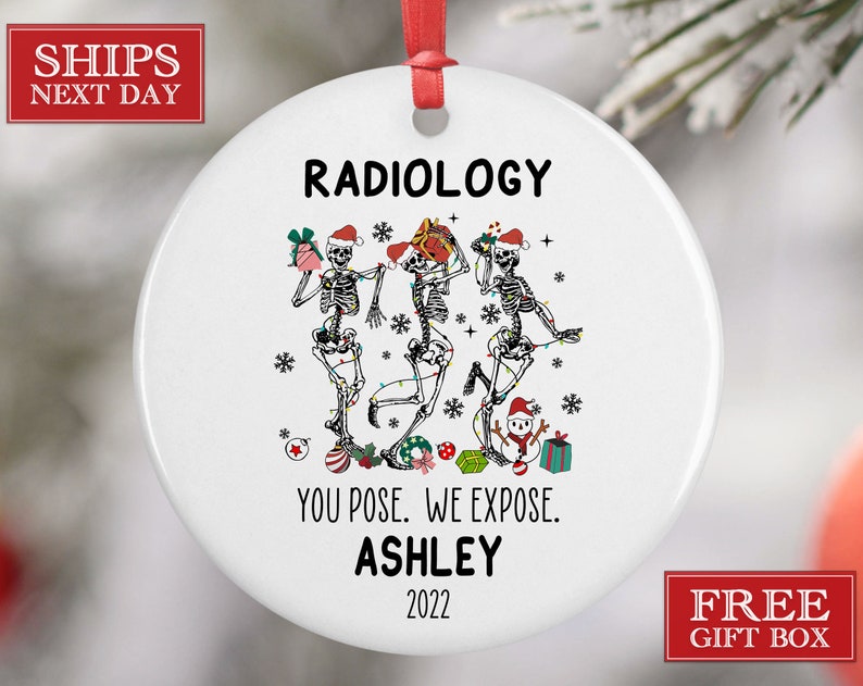 May include: White ceramic Christmas ornament with a red ribbon. The ornament features a cartoon image of three skeletons wearing Santa hats and holding gifts. The text "RADIOLOGY" is at the top of the ornament. Below the image is the text "YOU POSE. WE EXPOSE. ASHLEY 2022".