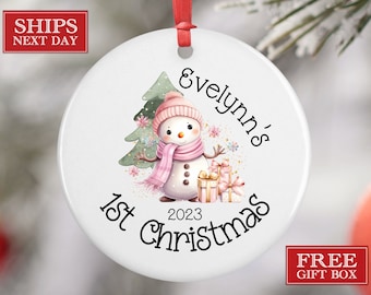 Może przedstawiać: Biała ceramiczna ozdoba choinkowa z ilustracją różowego i białego bałwanka, zielonego drzewa iglastego i tekstem "Evelyn's 1st Christmas 2023".