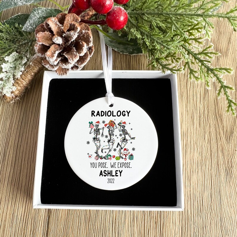 May include: A white round Christmas ornament with a black and white illustration of three skeletons wearing Santa hats and holding gifts. The ornament reads "Radiology You Pose. We Expose. Ashley 2022".