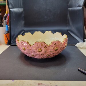 May include: A round ceramic bowl featuring a scalloped rim and a relief pattern of pink flowers with gold-painted centers and petal accents. The interior of the bowl is a smooth, solid cream color, while the exterior is covered in the floral design.