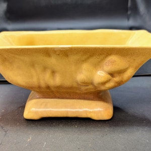 Puede incluir: Una maceta rectangular de cerámica con un esmalte brillante de color amarillo mostaza. El recipiente presenta un borde acampanado, un cuerpo cónico con sutiles detalles florales en relieve moldeados en los lados y una base de pedestal rectangular integrada.