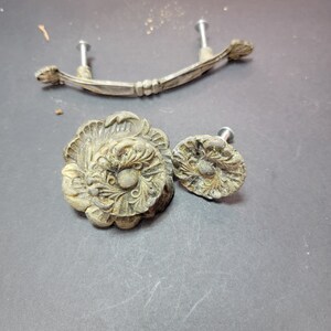 Puede incluir: Un juego de tres piezas de herraje metálico que incluye un tirador de cajón curvo con dos tornillos de montaje y dos pomos de armario redondos, todos con un acabado plateado desgastado y deslustrado con detalles florales en relieve.
