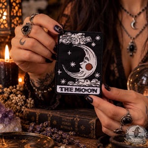 Peut inclure: Un écusson brodé rectangulaire représentant un croissant de lune blanc avec un visage, entouré d'étoiles et de nuages sur un fond noir. Le texte THE MOON est brodé en blanc au bas de l'écusson, qui est tenu par deux mains.