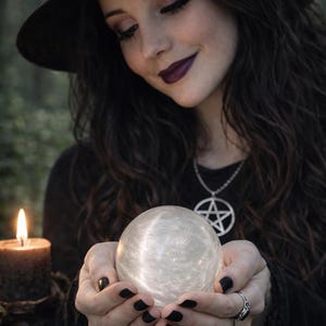 Op de afbeelding: Een vrouw met een zwarte hoed en donkere kleding houdt een gloeiende kristallen bol vast. Een brandende kaars en een pentagramketting zijn ook zichtbaar. Mystiek thema.