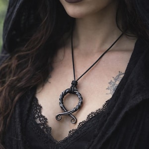Op de afbeelding: Een zwarte koordketting met een zilverkleurige hanger. De hanger heeft een cirkelvormig ontwerp met een gedraaide touwachtige textuur en een gestileerde, gebogen vorm aan de onderkant. De ketting wordt gedragen op een zwarte top.