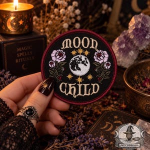 Puede incluir: Un parche bordado circular con fondo negro y borde rojo oscuro. El diseño presenta el texto "MOON CHILD" en crema, un gráfico central de luna blanca, estrellas doradas y dos arreglos florales con rosas de color rosa claro y hojas verdes.