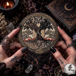Puede incluir: Un parche bordado circular que presenta un árbol central con raíces y ramas entrelazadas, flanqueado por dos figuras similares a lobos. El diseño está realizado en tonos oro, negro y crema, encerrado dentro de un borde decorativo trenzado.