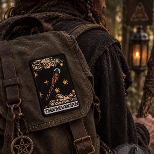 Op de afbeelding: Een rechthoekige geborduurde patch met een zwarte achtergrond, een bruine staf met een groene bol in het midden, omringd door gele sterren en witte wolken. De tekst THE MAGICIAN staat in het wit onderaan de patch.