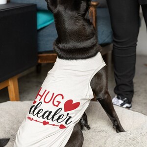 May include: A black dog wearing a white tank top with the text "hug dealer" and a red heart. The dog is sitting on a white fluffy rug.