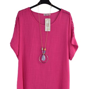 May include: A pink, short-sleeved, loose-fitting top with a round neckline and lace detailing on the shoulders. The top has a longer back hemline than the front. A gold necklace with a light blue teardrop pendant and colorful beads is worn with the top.