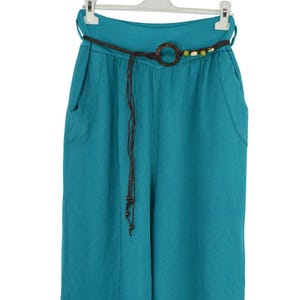 May include: Teal-colored wide-leg pants with a brown belt featuring a wooden ring and decorative beads. The pants have two front pockets and are hanging on a white hanger. The belt is made of a dark brown material.