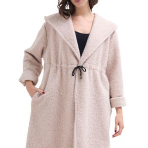 Op de afbeelding: Een beige cardigan met lange mouwen, een capuchon en een trekkoord in de taille. De cardigan heeft een zachte, pluche textuur.