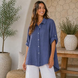 Puede incluir: Una camisa azul de manga larga con cuello, que presenta una tapeta frontal con botones visibles y un dobladillo trasero ligeramente más largo. La camisa tiene un corte relajado con mangas remangadas hasta los codos y tejido texturizado.