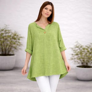 May include: A lime green tunic top with a V-neckline, a single brown button at the centre, and three-quarter length sleeves with rolled cuffs. The garment features a textured fabric finish and a loose, flowing silhouette paired with white trousers.