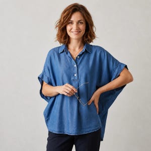May include: A blue denim-style short-sleeve shirt with a collar, a partial button-down front placket, and a loose, boxy fit. The shirt features a light blue wash, visible stitching along the seams, and a curved hemline that falls at the hips.